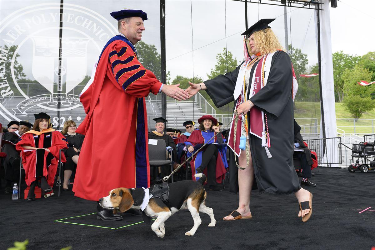 President Schmidt shakes hand with undergraduate female student crossing stage with her dog on a leash.