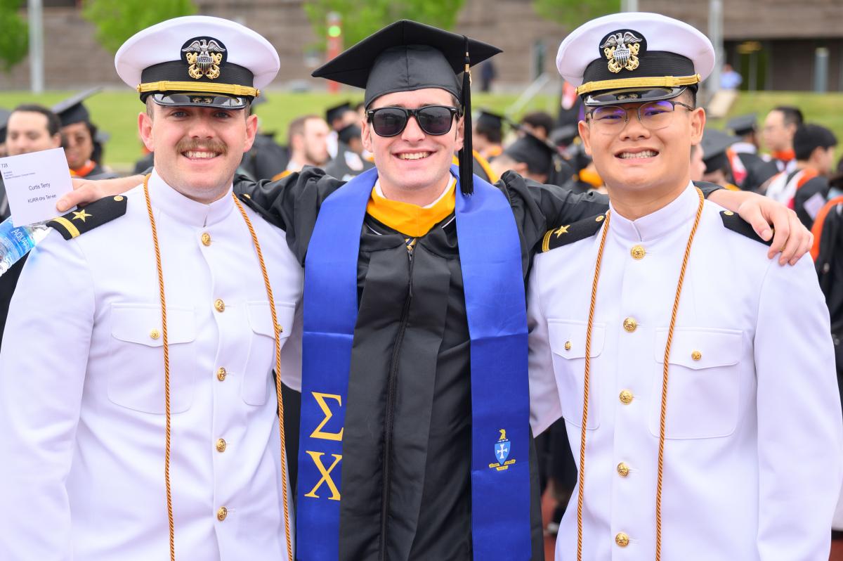 Three graduates posing for a photo at RPI's Commencement. The graduate in the middle is wearing a black cap and gown and sunglasses and the graduates on the left and right are wearing white naval uniforms and caps. 