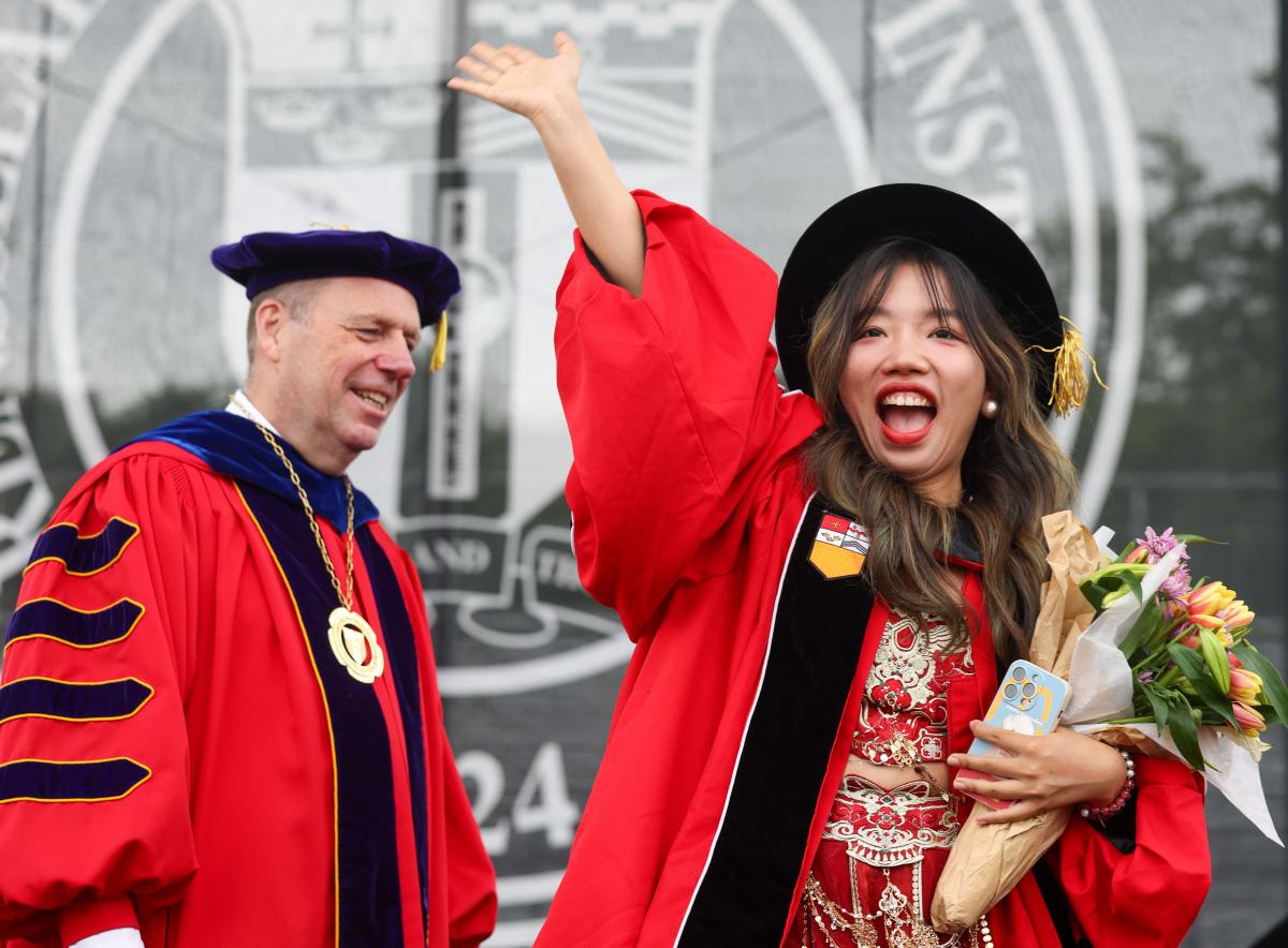 RPI doctoral graduate waves from the stage with President Martin Schmidt smiling in the background.