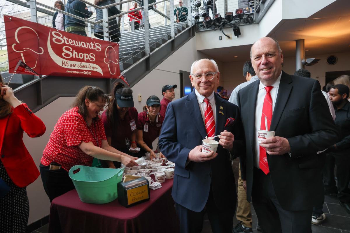 Congressman Paul Tonko and RPI President Martin Schmidt smiling and holding cups filled with ice cream from Stewart's Shops. Stewart's Shops sign in the background with people scooping additional cups of ice cream.