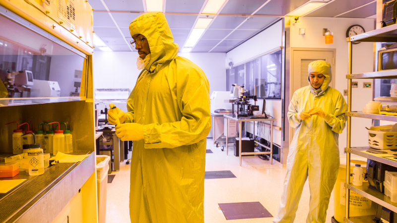 Two individuals in protective suits work in a laboratory with yellow lighting. One person is handling equipment near a counter, while the other is near the center of the room. Various lab equipment and containers are visible.
